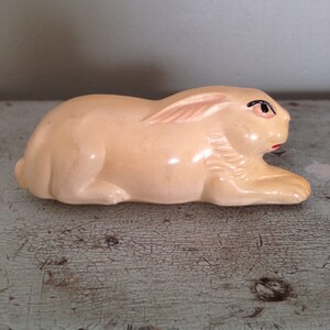 May include: A small, cream-colored ceramic rabbit figurine with black eyes and a red mouth. The rabbit is lying on its side with its head turned to the right.