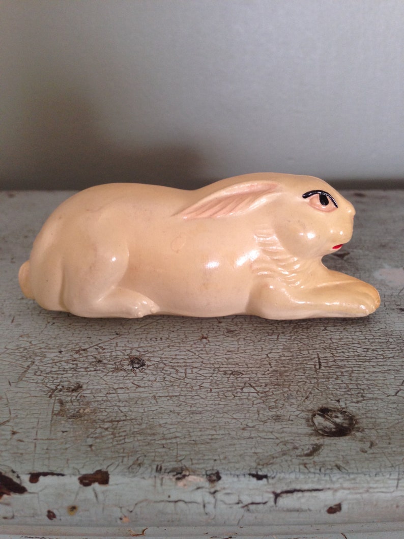 May include: A small, cream-colored ceramic rabbit figurine with black eyes and a red mouth. The rabbit is lying on its side with its head turned to the right.