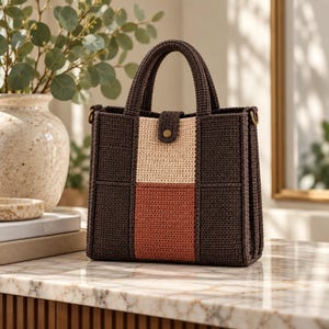 May include: A structured handbag crafted from dark brown crocheted yarn, featuring a vertical center panel with a cream-colored top section and a terracotta-colored bottom section. The bag includes two top handles and a small tab closure with a gold button.