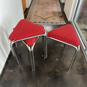 May include: Two stools with triangular seats upholstered in red fabric featuring a white grid pattern. Each stool has four slender, polished chrome metal legs that curve slightly outward at the base, providing a stable foundation for the seat.