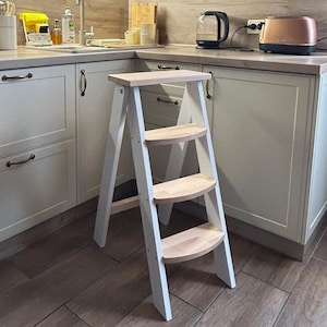 May include: A three-step folding ladder featuring white side frames and natural light-coloured wooden steps. The ladder has a flat top platform and three curved treads, constructed with a sturdy frame design for stability and reach in a kitchen.