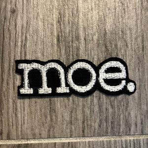 May include: A rectangular embroidered patch featuring the lowercase text "moe." in white thread against a solid black background. The lettering is rendered in a bold, serif-style font, and the entire patch is outlined with a thick black border.
