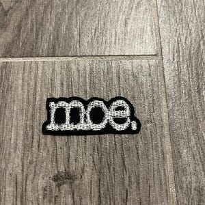 May include: A rectangular embroidered patch featuring the lowercase text "moe." in white thread against a solid black background. The letters are outlined in white, creating a bold contrast, and the patch has a distinct stitched border.