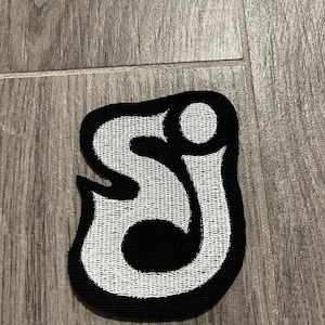 以下が含まれることがあります： スタイリッシュで連結されたフォントで「S」と「J」の文字が刺繍されたワッペン。黒い背景に白い糸で文字が形作られており、全体が太い黒の縁取りで囲まれているデザインです。