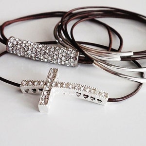 Puede incluir: Un brazalete de cuero marrón con un dije de cruz de plata y un dije de barra de diamantes de imitación de plata.