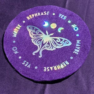 May include: A circular purple felt mat featuring a central holographic moth graphic, a moon phase icon, and the words REPHRASE, YES, NO, and MAYBE printed in a circular pattern around the edge in a reflective, iridescent holographic finish.