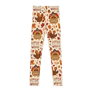 Op de afbeelding: Legging met volledige lengte op een gebroken witte achtergrond met een herhalend patroon van bruine kalkoenen met pelgrimshoeden, oranje en rode herfstbladeren, eikels en de tekst "GOBBLE TILL YOU WOBBLE" in bruine en oranje letters.