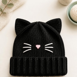 May include: A black knit beanie featuring two pointed cat ears at the top. The front of the hat is decorated with a small, embroidered pink heart-shaped nose and three white stitched whiskers on each side. The beanie has a thick ribbed cuff.