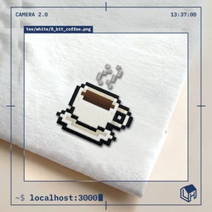 Op de afbeelding: Een wit t-shirt met een geborduurd 8-bit pixel art ontwerp van een koffiekopje op een schoteltje. Het kopje is wit met een donkerbruine vloeistof erin en een zwarte omlijning. Twee kleine grijze, gepixelde stoomwolkjes stijgen boven het kopje uit.