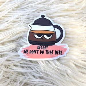 May include: A sticker featuring a cartoon coffee pot with an angry expression. The coffee pot is brown with a black handle and lid, and contains a brown liquid. The text "DECAF? WE DON'T DO THAT HERE." is below the pot.