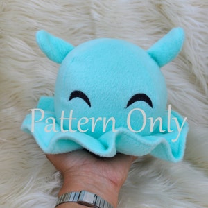 Puede incluir: Un juguete de peluche azul claro con forma de medusa con una cara sonriente. El juguete está siendo sostenido en una mano. El texto "Pattern Only" es visible en la imagen.