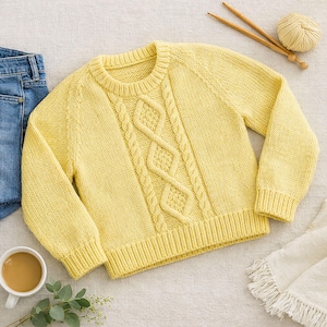 Può includere: Un maglione in maglia a maniche lunghe color giallo pallido con un motivo centrale a trecce verticali, scollo rotondo a coste e polsini a coste. Il maglione è mostrato insieme a un gomitolo di lana abbinato, due ferri da maglia e tessuto denim blu.