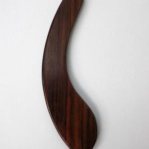 May include: A curved, dark brown wooden tool with a smooth, polished finish and visible wood grain patterns. The object features a tapered, elongated shape that narrows at one end and widens at the other, presenting a sleek, solid construction.