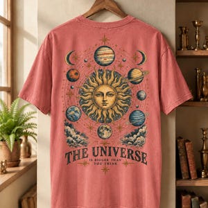 Könnte beinhalten: Ein korallenfarbenes T-Shirt mit kurzen Ärmeln und einem großen Grafikdruck auf der Rückseite. Das Motiv zeigt ein zentrales Sonnengesicht, umgeben von verschiedenen Planeten, Sternen und Wolken. Unter der Illustration steht der Text: THE UNIVERSE IS BIGGER THAN YOU THINK.