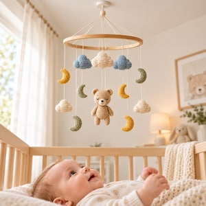 May include: A hanging nursery mobile featuring a circular wooden frame with suspended crochet elements including a central beige bear, blue and cream clouds, and yellow and sage green crescent moons, all attached by white strings.