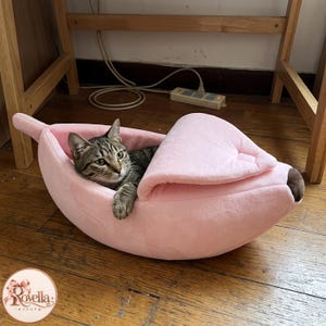Pode incluir: Uma cama de pelúcia para animais de estimação em rosa claro, com formato de banana descascada e ponta marrom. Um gato malhado marrom de olhos verdes descansa dentro da estrutura curva e aberta, com uma pata apoiada na borda do tecido.