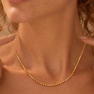 Peut inclure: Un collier doré composé d'une chaîne fine alternant de petites perles rondes et des entretoises cylindriques allongées. Ce bijou métallique repose sur le cou, mettant en valeur un motif répétitif de perles et de barres à la finition polie.