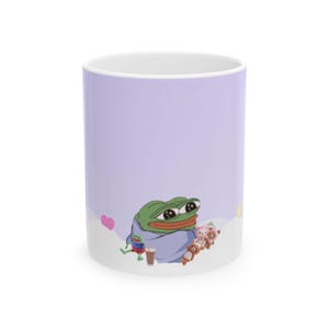 May include: A light purple ceramic mug featuring a graphic of a green frog character wrapped in a blue blanket, holding a brown drink, accompanied by three small brown and pink bear figures, a small pink heart, and white cloud shapes.