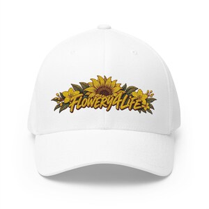 May include: A white baseball cap featuring an embroidered design on the front. The embroidery depicts a large yellow sunflower flanked by smaller yellow flowers and green leaves, with the text FLOWERY4LIFE in a bold, yellow, stylized font.
