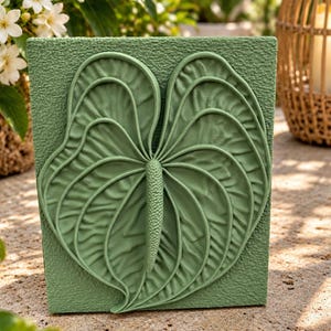 Peut inclure: Une plaque murale rectangulaire vert sauge présentant un relief tridimensionnel d'une feuille d'anthurium et de son spadice. Le fond de la plaque possède une surface texturée et granuleuse, tandis que le motif de la feuille montre des nervures.