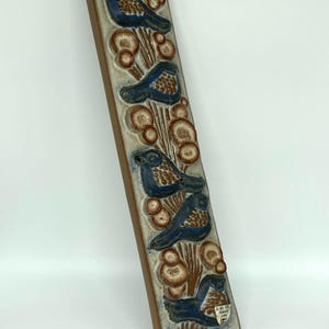 Könnte beinhalten: Eine rechteckige Wandplatte aus Keramik mit einem Reliefdesign aus fünf blauen Vögeln, die zwischen braunen Kiefernnadeln und runden Kiefernzapfen sitzen. Das Stück hat einen cremefarbenen Hintergrund mit blauen Sprenkeln und einen kleinen weißen Aufkleber.