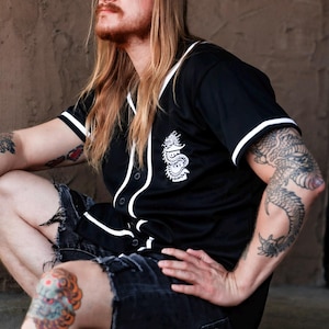 May include: A black short-sleeved button-down baseball jersey featuring white piping along the collar, front placket, and sleeve cuffs. A white graphic of a dragon coiled around a pagoda is printed on the left chest area of the shirt.
