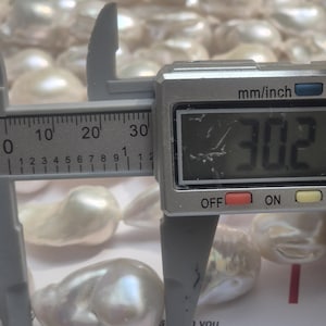 Puede incluir: Primer plano de perlas blancas brillantes de forma irregular. Un calibrador digital mide una perla a 3,02 cm. Las perlas tienen una superficie lisa e iridiscente, con un color suave y cremoso. El fondo muestra una colecci&oacute;n de perlas similares.