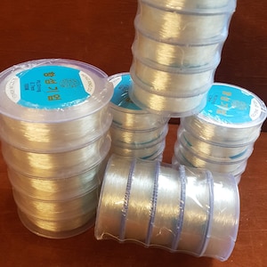 Puede incluir: Varias bobinas de hilo elástico transparente, cada una envuelta en plástico y etiquetada con el texto "Elastic String 0.8mm 100M".