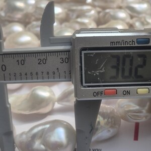 Puede incluir: Primer plano de perlas blancas lustrosas de forma irregular. Un calibrador digital mide una perla a 3,02 cm. Las perlas est&aacute;n sobre una superficie blanca, con texto visible en el fondo.