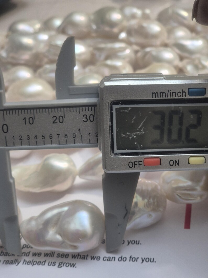 Puede incluir: Primer plano de perlas blancas lustrosas de forma irregular. Un calibrador digital mide una perla a 3,02 cm. Las perlas est&aacute;n sobre una superficie blanca, con texto visible en el fondo.