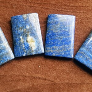 May include: Four rectangular lapis lazuli stones with gold flecks. The stones are arranged in a row on a brown surface.