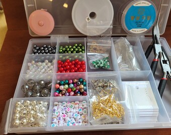 Può includere: Una scatola organizer in plastica trasparente riempita con forniture per bigiotteria, tra cui perline di vari colori, perline con lettere, ganci dorati per orecchini, un metro a nastro rosa, una bobina di filo elastico blu e pinze.