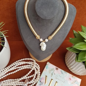 Puede incluir: Un collar dorado con un colgante de perla grande e irregular y dos perlas m&aacute;s peque&ntilde;as. El collar se exhibe sobre un busto gris, con un collar de perlas y una tarjeta de felicitaci&oacute;n que dice "To a Special Mum" cerca.