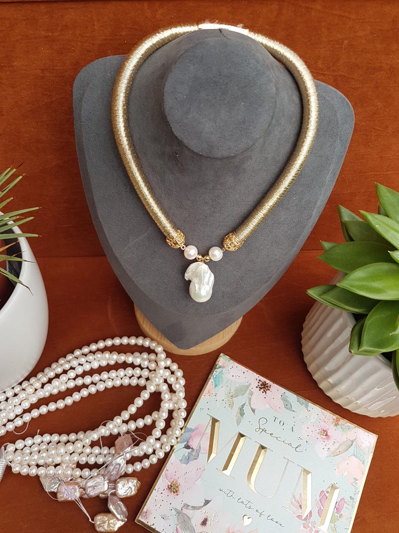 Puede incluir: Un collar dorado con un colgante de perla grande e irregular y dos perlas m&aacute;s peque&ntilde;as. El collar se exhibe sobre un busto gris, con un collar de perlas y una tarjeta de felicitaci&oacute;n que dice "To a Special Mum" cerca.