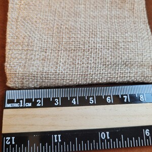 May include: Close-up of a beige fabric with a woven texture. A ruler is placed below the fabric, showing measurements in inches and centimeters.