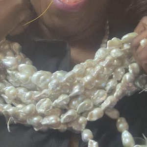 Puede incluir: Collar de perlas de m&uacute;ltiples hilos con perlas grandes de forma irregular. Las perlas son de color blanco cremoso y tienen un brillo lustroso. Un anillo con una perla es visible en una mano que sostiene el collar.