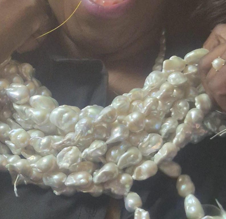 Puede incluir: Collar de perlas de m&uacute;ltiples hilos con perlas grandes de forma irregular. Las perlas son de color blanco cremoso y tienen un brillo lustroso. Un anillo con una perla es visible en una mano que sostiene el collar.