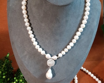 Pode incluir: Um colar de pérolas brancas com um grande pingente de pérola branca e um fecho de prata. O colar está em um busto de manequim cinza. Há um segundo colar de pérolas mais curto e um par de brincos de pérolas na mesa abaixo do busto.