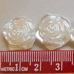 Pode incluir: Um close-up de uma guarnição de rosa de pérola branca. A guarnição tem aproximadamente 1 cm de largura e apresenta um padrão repetitivo de rosas tridimensionais. As rosas são feitas de madrepérola branca e têm um brilho ligeiramente iridescente.