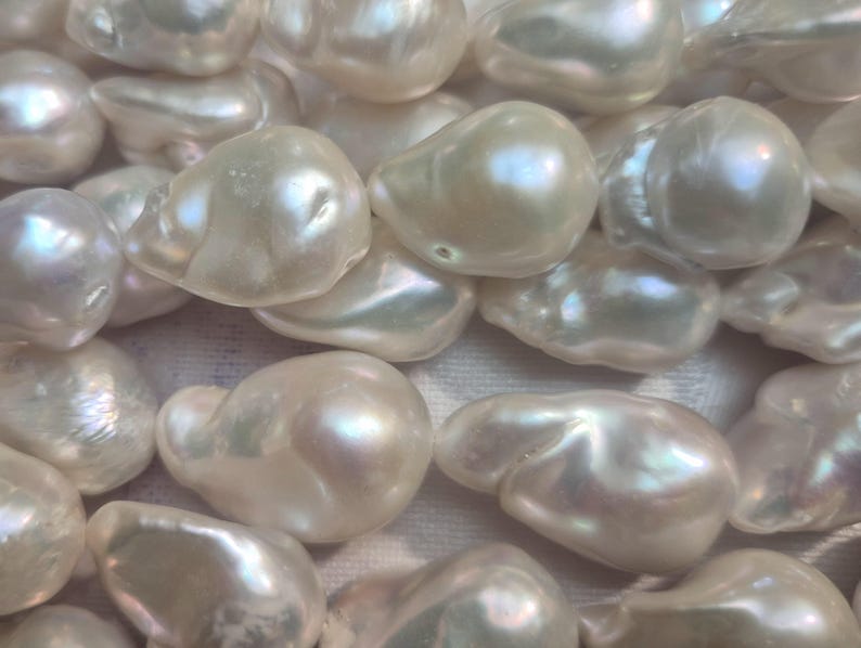 Puede incluir: Primer plano de una colecci&oacute;n de perlas lustrosas de forma irregular. Las perlas tienen un color blanco cremoso con reflejos iridiscentes, que reflejan la luz. Las perlas est&aacute;n agrupadas, mostrando sus formas org&aacute;nicas &uacute;nicas.