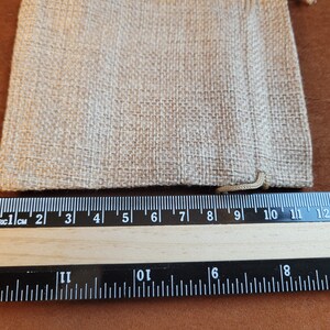 May include: A close-up of a natural beige burlap drawstring bag measuring 9 inches wide. The bag is lying flat on a brown surface next to a ruler.