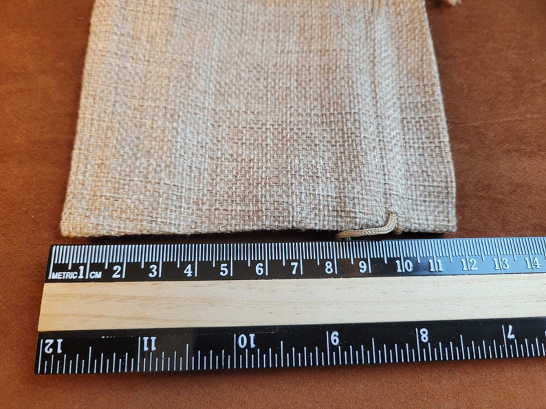 May include: A close-up of a natural beige burlap drawstring bag measuring 9 inches wide. The bag is lying flat on a brown surface next to a ruler.