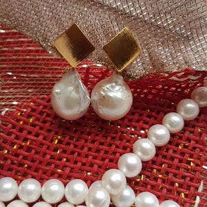 Puede incluir: Pendientes de perlas con cuadrados dorados y perlas blancas grandes de forma irregular. Un collar de perlas blancas sobre una superficie tejida roja, con un elemento decorativo dorado y plateado en el fondo.