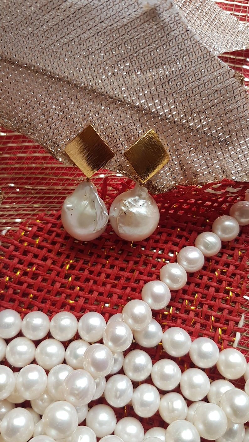 Puede incluir: Pendientes de perlas con cuadrados dorados y perlas blancas grandes de forma irregular. Un collar de perlas blancas sobre una superficie tejida roja, con un elemento decorativo dorado y plateado en el fondo.
