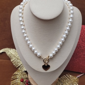 Könnte beinhalten: Eine Perlenkette mit einem goldenen Herzanhänger. Die Kette wird auf einer cremefarbenen Büste präsentiert. Die Perlen sind weiß und rund. Der Herzanhänger ist goldfarben und hat eine glatte Oberfläche. Die Halskette ist ein klassisches Schmuckstück.