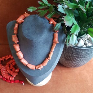 Puede incluir: Un collar de coral hecho de cuentas de color naranja sobre un expositor de terciopelo negro. El collar está al lado de un montón más pequeño de cuentas de coral.