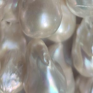 Puede incluir: Primer plano de una colecci&oacute;n de perlas blancas lustrosas de forma irregular. Las perlas tienen una superficie lisa e iridiscente con sutiles toques de color, que reflejan la luz. Las perlas est&aacute;n ensartadas, lo que sugiere que son para la fabricaci&oacute;n de joyas.