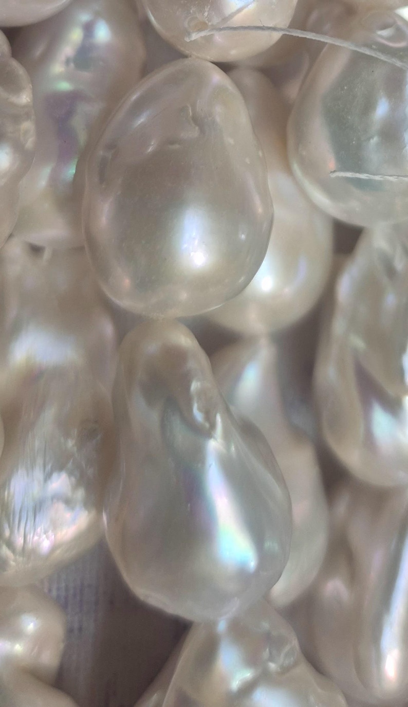 Puede incluir: Primer plano de una colecci&oacute;n de perlas blancas lustrosas de forma irregular. Las perlas tienen una superficie lisa e iridiscente con sutiles toques de color, que reflejan la luz. Las perlas est&aacute;n ensartadas, lo que sugiere que son para la fabricaci&oacute;n de joyas.