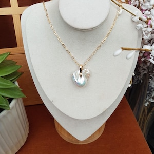 Puede incluir: Un collar de cadena dorada con un colgante de perla iridiscente de forma irregular. El collar se exhibe sobre un busto blanco, sobre un fondo marr&oacute;n c&aacute;lido, con vegetaci&oacute;n y flores en la periferia.