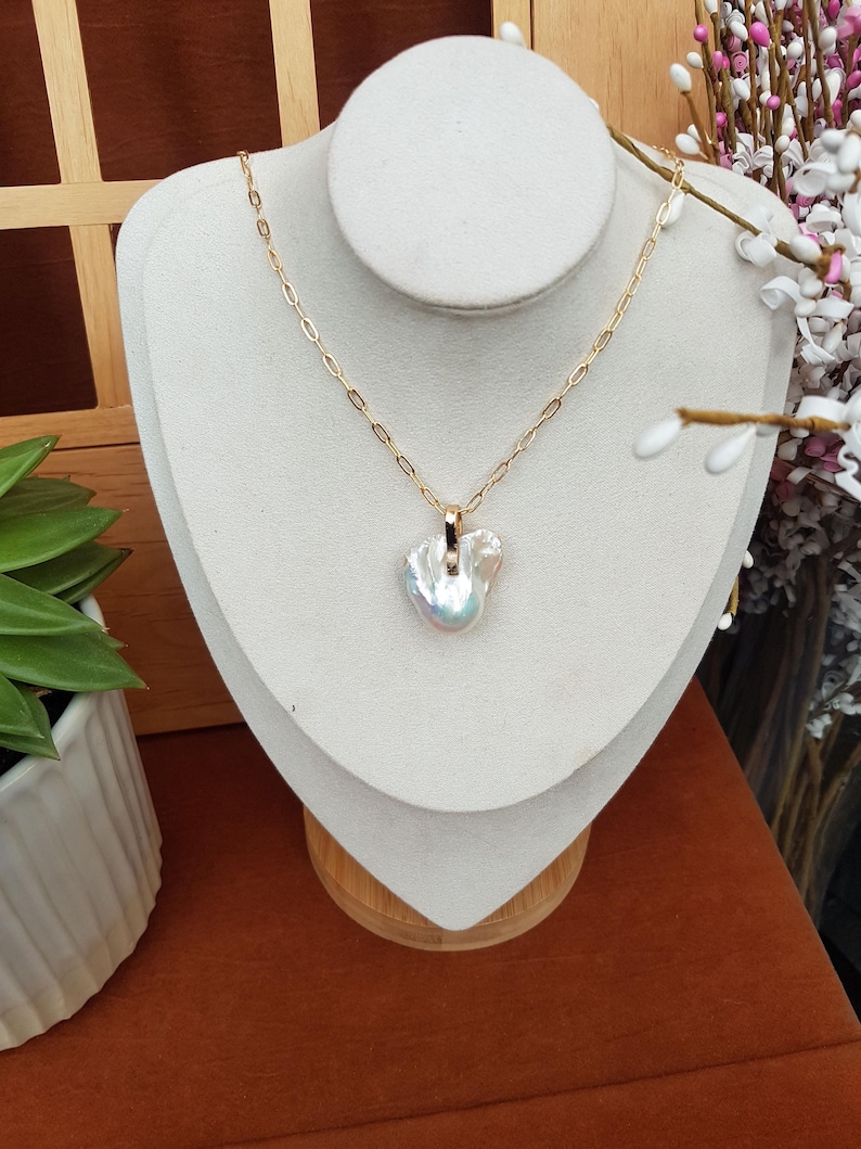 Puede incluir: Un collar de cadena dorada con un colgante de perla iridiscente de forma irregular. El collar se exhibe sobre un busto blanco, sobre un fondo marr&oacute;n c&aacute;lido, con vegetaci&oacute;n y flores en la periferia.